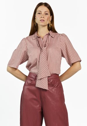 Vrouw met lang bruin haar die gouden oorbellen draagt, een roze gestreept kort mouw blouse met een strik aan de voorkant, en hoge taille rode leren broek.