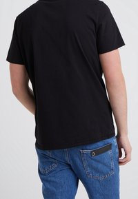 Camiseta de algodón negra de manga corta, vista desde la parte de atrás. Combinada con jeans de mezclilla azul que presentan un parche negro en el bolsillo trasero.