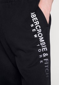 Pantalones de chándal negros con una textura suave, que cuentan con un logo impreso en blanco en el muslo izquierdo que dice “ABERCROMBIE & FITCH NEW YORK.”