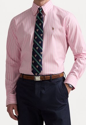 Mężczyzna w różowo-białej pasiastokoszuli na guziki, granatowym krawacie w wzory, brązowym skórzanym pasku i granatowych spodniach z jedną ręką w kieszeni.
