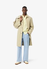 Cappotto beige, camicia a righe gialle, jeans a gamba larga blu chiaro, sneakers beige e tan. Modello che tiene una borsa, in piedi su uno sfondo bianco.