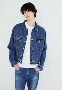 Denim jakna z dvema sprednjima žepoma, z gumbom, v bledomodri barvi; nošena čez belo srajco, v paru z usklajenimi kavbojkami.