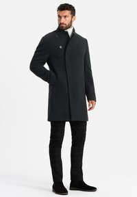 Abrigo de lana gris oscuro con un cuello alto, cierre de dos botones y bolsillos laterales. Combinado con pantalones a cuadros negros y botas negras de tobillo.