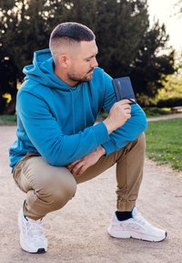 Black textured wallet held by a person wearing a blue hoodie and beige pants, outdoors with greenery in the background.