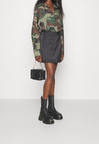 Donna che indossa una camicia a maniche lunghe camouflage, una mini gonna nera, stivali neri chunky e tiene una piccola borsa nera con una tracolla a catena.