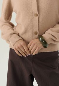 Mani con unghie rosa chiaro incrociate sopra pantaloni scuri, indossando un maglione beige con bottoni e un bracciale spesso verde scuro al polso.