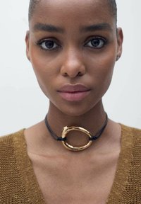 Collier ras du cou présentant un cercle en métal doré poli sur un cordon noir. Le design dispose de textures lisses et d'une forme circulaire audacieuse.