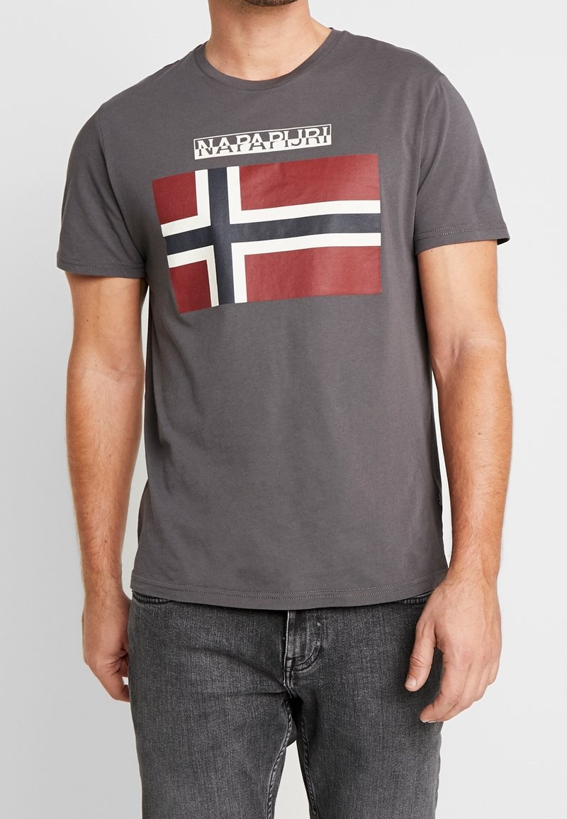 Homme portant un t-shirt gris avec un drapeau croix de style norvégien rouge, blanc et bleu et le texte « NAPAPIJRI » imprimé sur la poitrine.