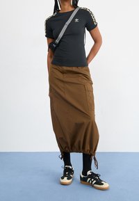 Person som bär en svart Adidas-tröja med logotyp, en brun midi-kjol med dragsko nertill, svarta strumpor och svartvita sneakers, stående på blått golv.