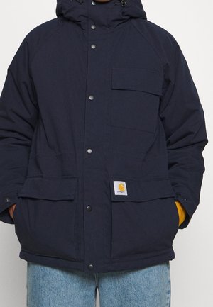 Personne portant une veste à capuche Carhartt bleu marine avec boutons pression et plusieurs poches, associée à un jean bleu clair.