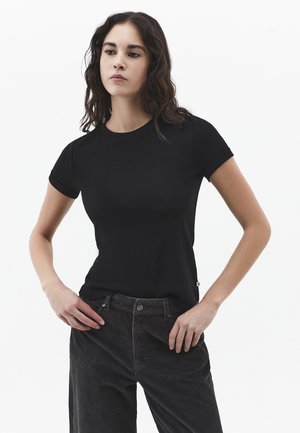 Joven con cabello largo y oscuro, vestida con una camiseta negra ajustada de manga corta y pantalones oscuros de pana, de pie con las manos en las caderas.