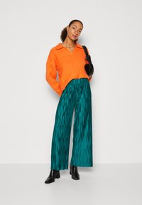 Jersey de color naranja brillante con cuello, combinado con pantalones de pierna ancha de color teal y con patrón, y botas negras hasta el tobillo. La modelo sostiene un bolso negro.