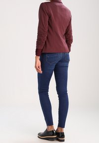 Personne portant une veste moulante bordeaux, un jean skinny bleu foncé et des mocassins noirs brillants, debout de dos face à la caméra.