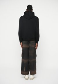 Μαύρη κουκούλα με κορδόνι που συνδυάζεται με χαλαρούς παντελόνες τύπου cargo με κεκλιμένο μοτίβο σε σκούρο καφέ και μαύρο χρώμα. Λευκά αθλητικά παπούτσια.