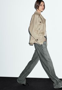 Trench coat beige doppiopetto con grandi bottoni, abbinato a jeans in denim grigio a taglio largo. Texture liscia e silhouette rilassata.