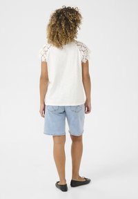 Weißes kurzärmliges Oberteil mit Spitzenverarbeitung an den Schultern, kombiniert mit hellblauen Denim-Shorts und schwarzen Ballerinas, von hinten betrachtet.