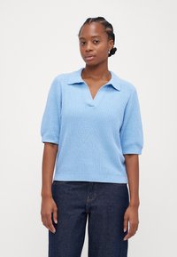 Suéter tricotado azul claro com gola polo, mangas curtas e um acabamento texturizado, combinado com calças de ganga escuras.