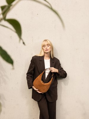 Donna bionda con un completo marrone scuro tiene una borsa in camoscio color sabbia contro una parete beige uniforme, con foglie verdi sfocate in primo piano.