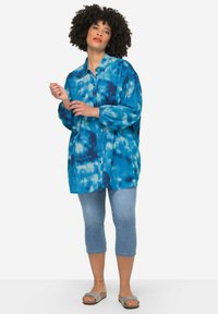 Camicetta a maniche lunghe in tie-dye blu, realizzata in tessuto leggero. Indossata con jeans alla caviglia e sandali argentati.