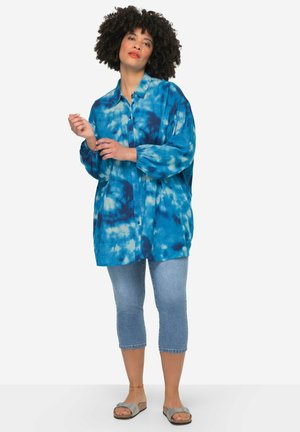 Chemise boutonnée bleu tie-dye à manches longues, en tissu léger. Portée avec un jeans cropped et des sandales en argent.