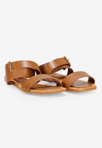 Sandalias de cuero marrón con dos correas anchas y suela plana. Con una hebilla metálica en una de las correas. Textura suave con un diseño minimalista.
