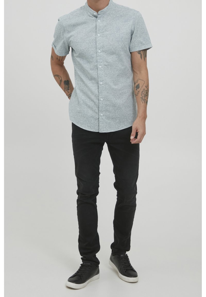 Homme portant une chemise grise à manches courtes col mandarin, pantalon noir ajusté et baskets noires, debout les mains détendues. Tatouages visibles sur les bras.
