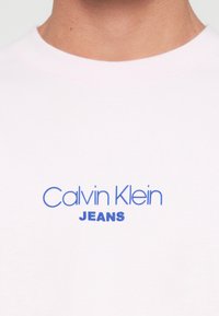 Nahaufnahme eines weißen Hemdkragens mit blauer "Calvin Klein JEANS"-Aufschrift in der Mitte vorne.