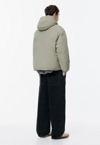 Olivovo zelená prešívaná bunda s kapucňou, vyrobená z ľahkého materiálu, štvorcový strih a prešité detaily, nosená cez čierno-biele kárované tričko.