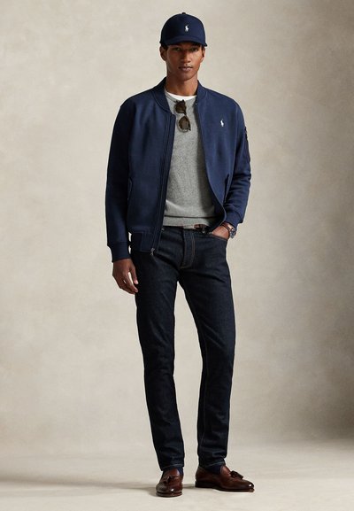 Homme portant une casquette marine, une veste bomber marine, un pull gris, un jean foncé, des mocassins marron à glands et des lunettes de soleil suspendues à l'encolure du pull.