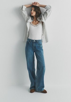 Naine seisab käed üles tõstetud tühja seina ees, seljas triibuline bleiser, valge pitsist top, laiade säärtega teksad ja pruunid jalanõud.