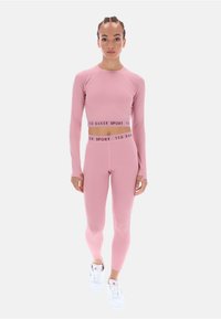 Rózsaszín, hosszú ujjú sportfelső crop fazonban, hozzá illő rózsaszín leggings és fehér tornacipő, amelynek derékpántján merész logó található.