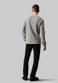 Camisa de manga larga gris claro, texturizada y con cuello redondo, combinada con pantalones negros. Presenta un sutil patrón acanalado y puños ajustados.