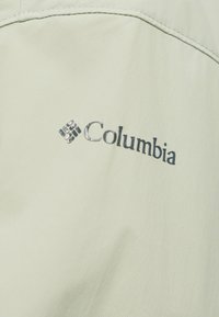 Λογότυπο Columbia σε μαύρο χρώμα πάνω σε ανοιχτό μπεζ ύφασμα με ορατές ραφές κοντά στην κορυφή του υφάσματος.