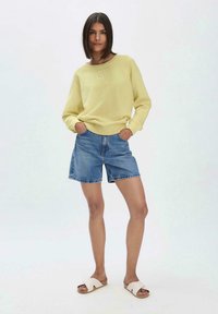 Gelbes Sweatshirt mit floralen Stickereien, kombiniert mit blauen Jeansshorts und hellen Sandalen, vor einem neutralen Hintergrund präsentiert.