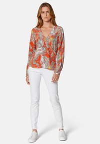 Rote Bluse mit Paisley-Muster, tiefem V-Ausschnitt, langen Ärmeln und lockerer Passform, kombiniert mit weißen Slim-Fit-Jeans und weißen Sneakers.