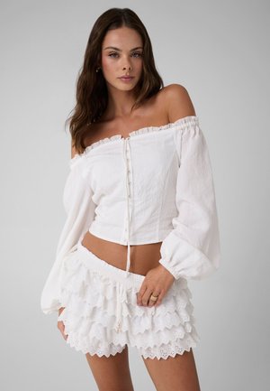Femme portant un chemisier blanc à manches longues décolleté aux épaules et un short blanc à volants superposés, posant devant un fond gris uni.