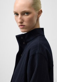 Camicia blu navy con tessuto strutturato, colletto alto e dettagli con bottoni sul fronte. Il design presenta sottili righe verticali.