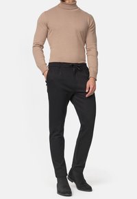 Mężczyzna w beżowym golfie, czarnych spodniach z troczkami oraz czarnych zamszowych butach za kostkę, stoi z jedną ręką w kieszeni, na białym tle.