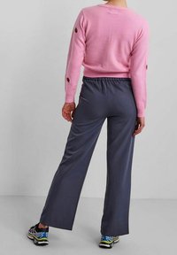 Person som bär en rosa tröja med jordgubbsapplikationer, lösa mörkgrå byxor och flerfärgade träningsskor, stående mot en enfärgad grå bakgrund.