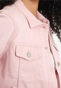 Chaqueta de mezclilla rosa con cuello, cierre de botones y dos bolsillos frontales con acentos metálicos. La textura es suave con detalles de costura visibles.