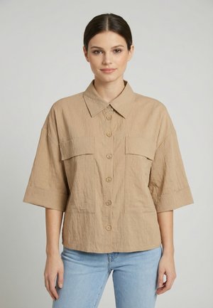 Vrouw draagt een beige overhemd met grote voorzakken en ellebooglange mouwen, gecombineerd met lichtblauwe spijkerbroek, staand tegen een effen achtergrond.