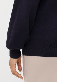 Pulôver azul-marinho com um corte descontraído, mangas volumosas e punhos em canelado; usado sobre um tecido bege claro, destacando a suavidade da textura e o contraste de cores.