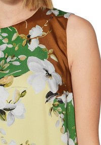 Ärmellose Bluse mit floralem Muster, weißen Blumen und grünen Blättern auf gelbem Hintergrund, ergänzt durch einen braunen Abschnitt.