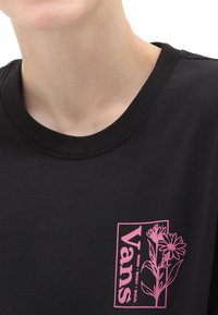 T-shirt in cotone nero con un logo floreale rosa sul lato sinistro del petto e testo. Design semplice con collo rotondo, texture morbida e linee pulite.