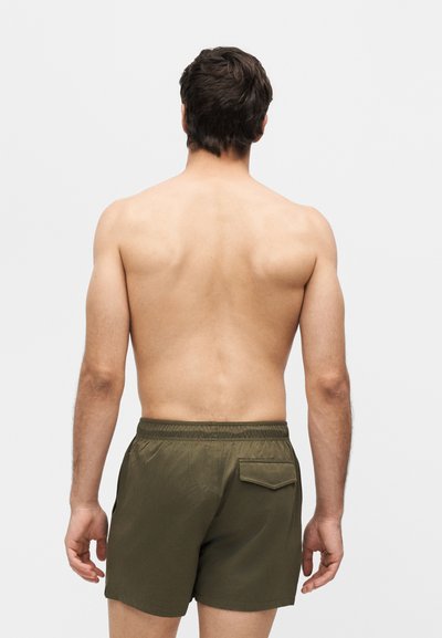 Άνδρας με κοντά σκούρα μαλλιά, χωρίς πουκάμισο, φοράει χακί σορτς με πίσω τσέπη, γυρισμένος πλάτη σε απλό φωτεινό φόντο.