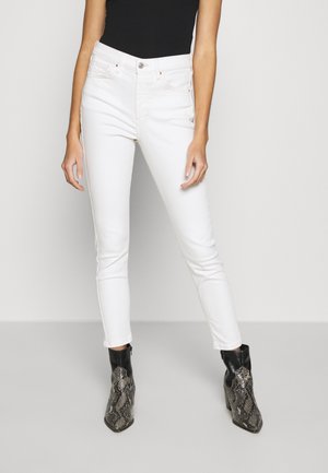 Jeans blancos ajustados con una cintura alta y detalles de costura sutiles, combinados con botas negras de tobillo estampadas y una blusa negra ajustada.