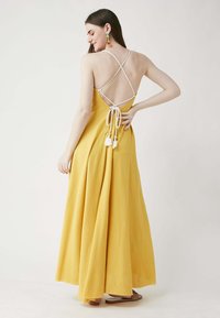 Maxikleid aus sonnengelbem Stoff mit fließender Silhouette, offener Rückenpartie, weißen, gekreuzten Trägern und Quastenakzenten an der Taille.