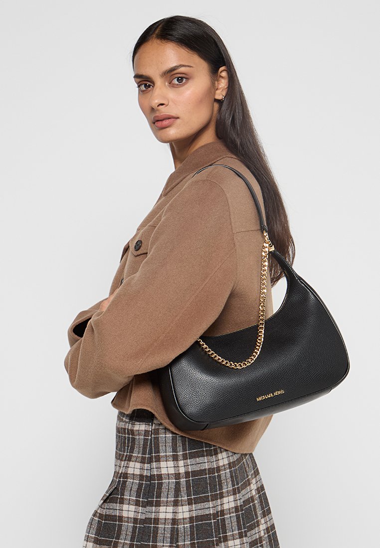 Borsa a spalla in pelle nera con finitura textured, accentata da catena dorata e dettaglio del logo, abbinata a una giacca marrone e una gonna a quadri.