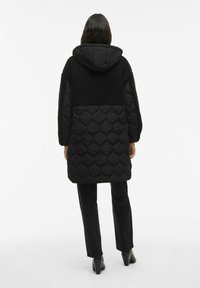 Sieviete, tērpusies līdz ceļgaliem garā melnā kvaltētajā un flisa mēta ar kapuci, melnās biksēs un melnās augstpapēžu potīšu zābakos, redzama no aizmugures.