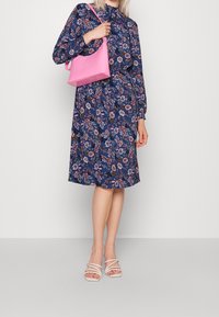 Robe à imprimé floral bleu marine avec manches longues, taille cintrée et col à volants. Associée à un sac à main rose et des talons blancs à brides.